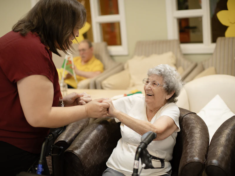 A caregiver holding hands with a smiling older woman as she gets ready to stand from her armchair, her walker beside her