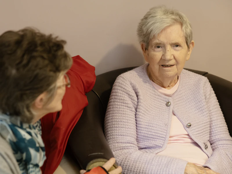 An older woman in a lilac cardigan chatting with a visitor on a sofa